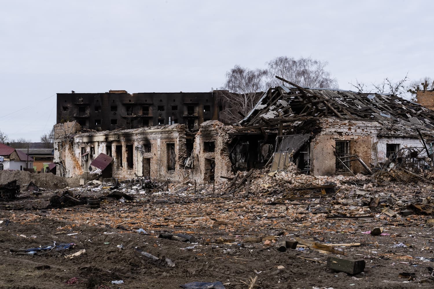 Разрушенное кирпичное здание без крыши и с выгоревшими окнами на фоне обугленного многоэтажного дома. На переднем плане — груды обломков и мусора.
