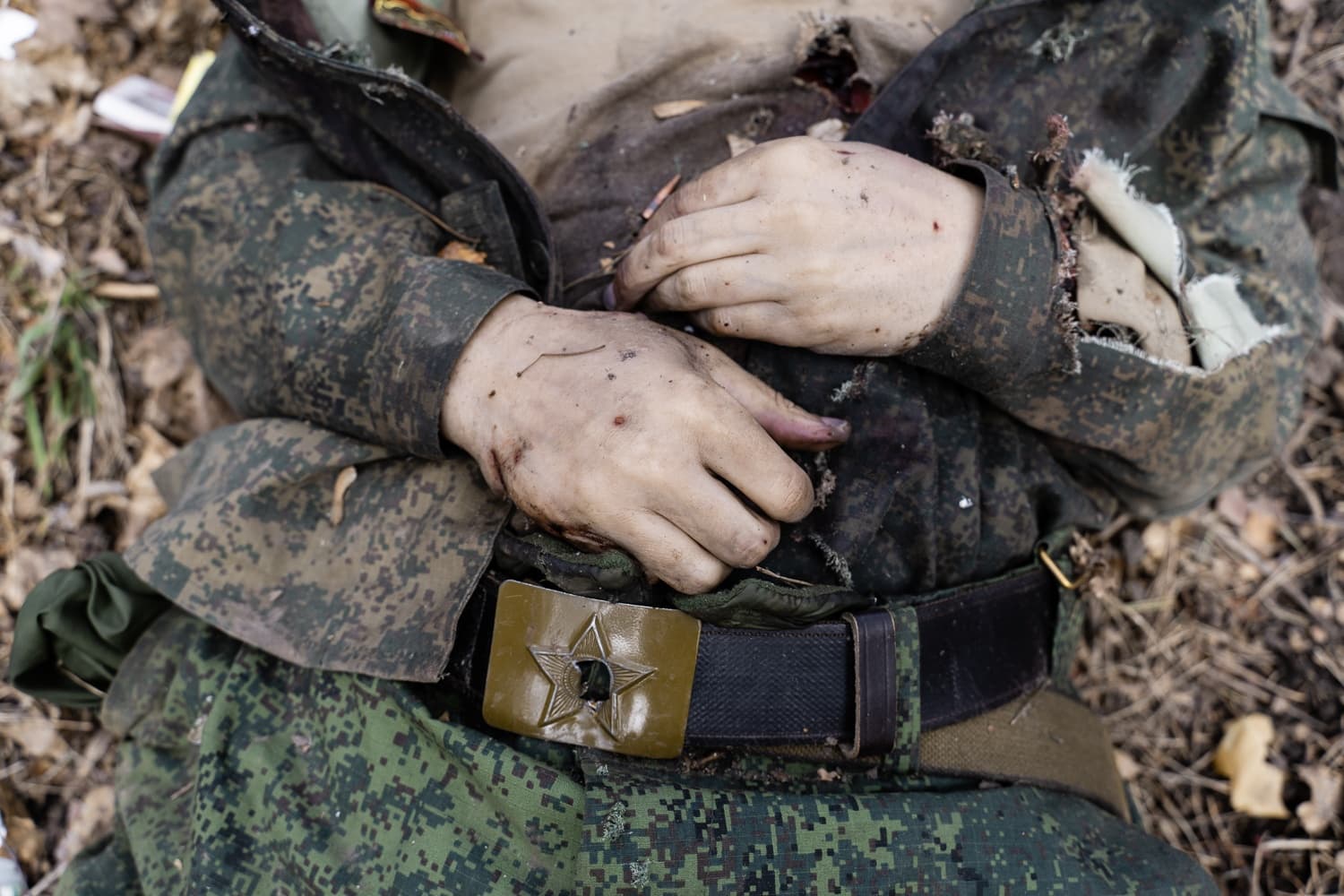 Тело погибшего солдата в камуфляже, лежащее на земле. Видны руки, поврежденная униформа и армейский ремень с пряжкой-звездой.