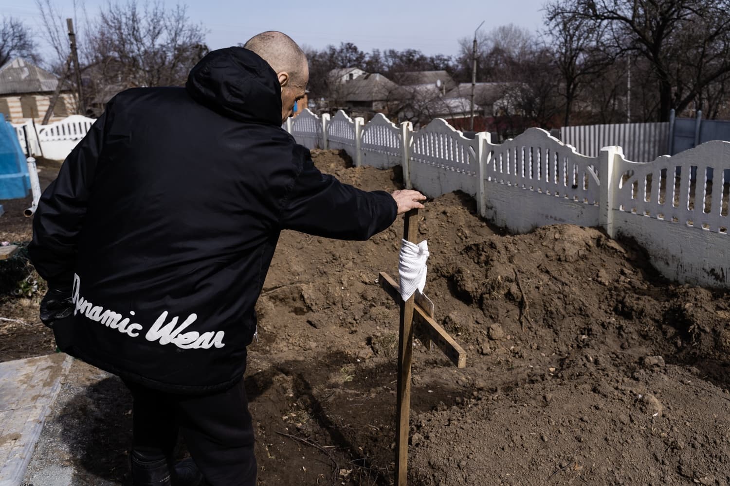 Мужчина в куртке "Dynamic Wear" склонился над свежей могилой, касаясь простого деревянного креста.