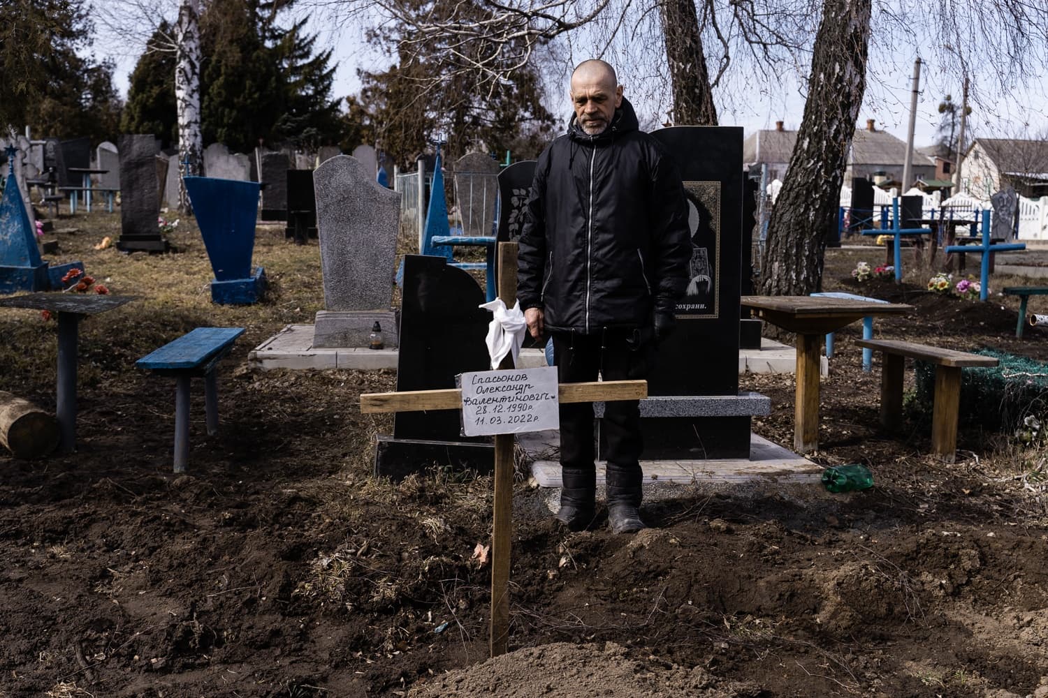Мужчина в черном стоит перед свежей могилой на кладбище, отмеченной деревянным крестом. На кресте: "ЕПЛАСОНОВ АЛЕКСАНДР ВАЛЕНТИНОВИЧ 28.12.1990 — 11.03.2022Г".