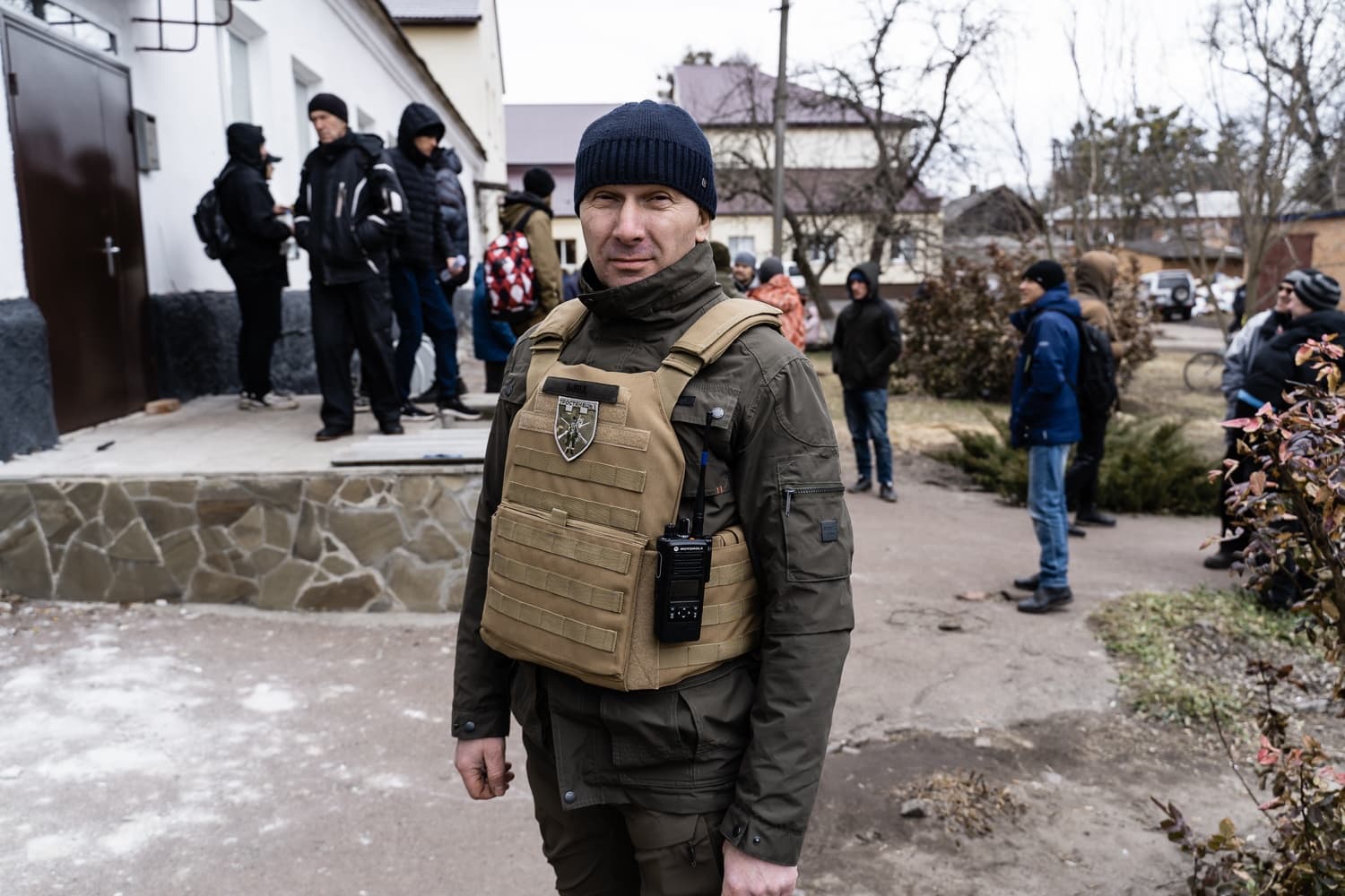 Мужчина в тактической одежде, шапке и бежевом бронежилете с рацией стоит на улице. На заднем плане видна группа гражданских лиц.