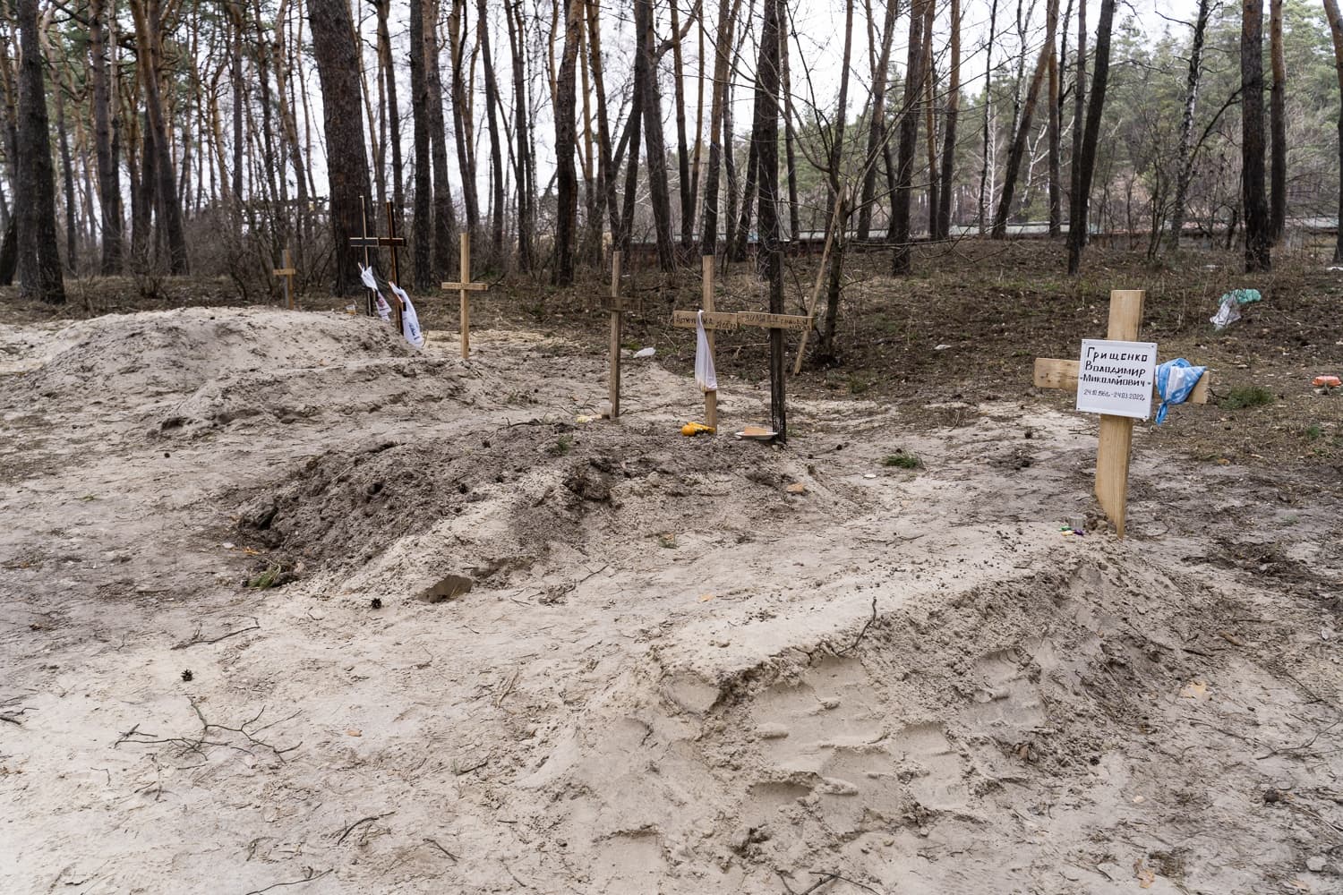 Ряд свежих насыпей в лесу, отмеченных простыми деревянными крестами. На ближнем кресте табличка: "Грищенко Володимир Миколайович - 24.11.1944- 24.03.2022".