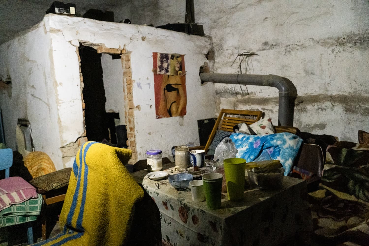 Сырой подвал, используемый как убежище, со столом, заваленным чашками, и кирпичной пристройкой с отверстием вместо двери. На стене висит плакат с изображением женщины в бикини.