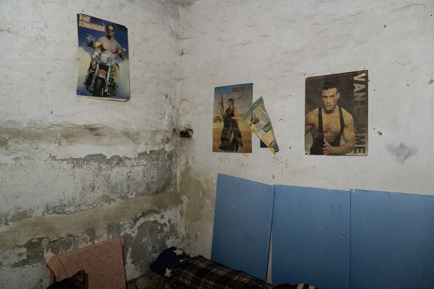 Угол ветхого помещения. На стене постеры: «Arnold SCHWARZENEGGER» на мотоцикле и «VAN DAMME». Внизу синие панели и спальное место.
