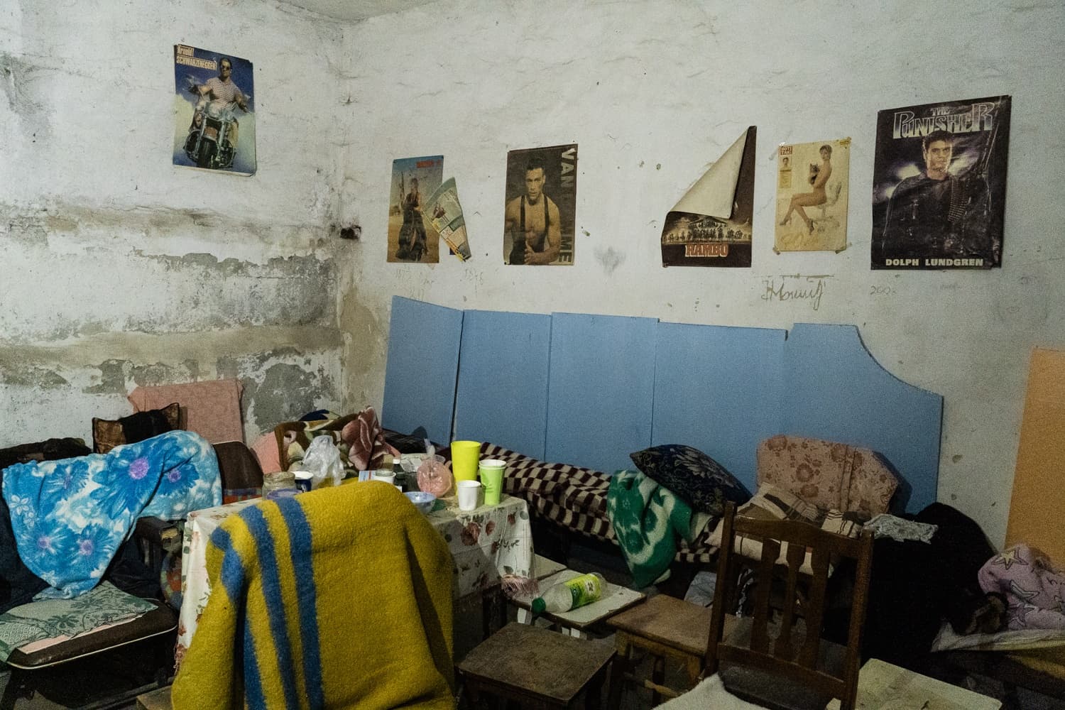 Обшарпанная комната с импровизированными спальными местами и разбросанными одеялами. Стены увешаны старыми кинопостерами: Шварценеггер на мотоцикле, "VAN DAMME", "RAMBO", и "THE PUNISHER" с "DOLPH LUNDGREN".