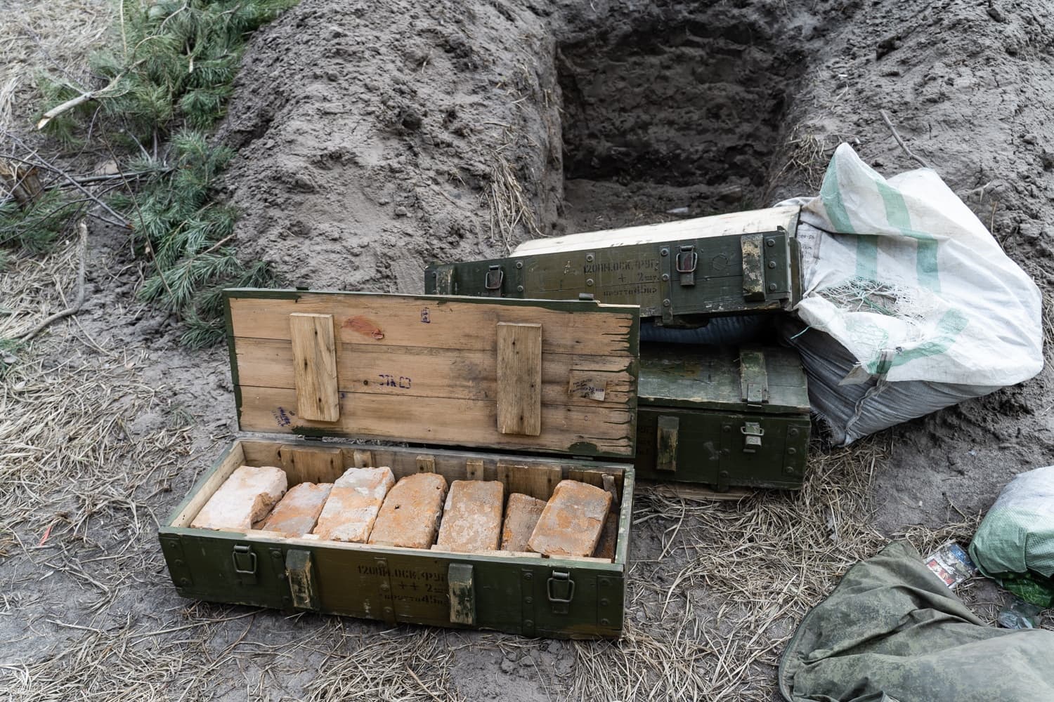 Открытый военный ящик для боеприпасов, с маркировкой, включающей "120мм ОФС", заполнен кирпичами. Рядом лежат другие ящики и насыпь земли.