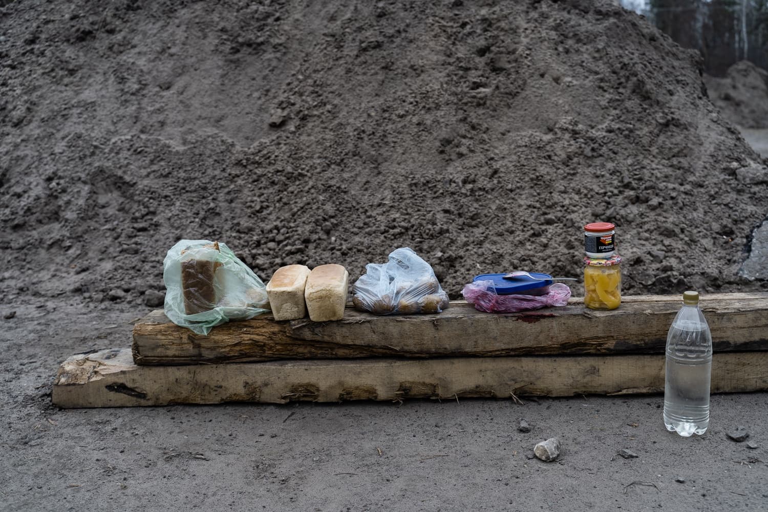 Хлеб, овощи, консервы, банка с "САЛО" и бутылка воды, разложенные на деревянных досках перед насыпью земли.