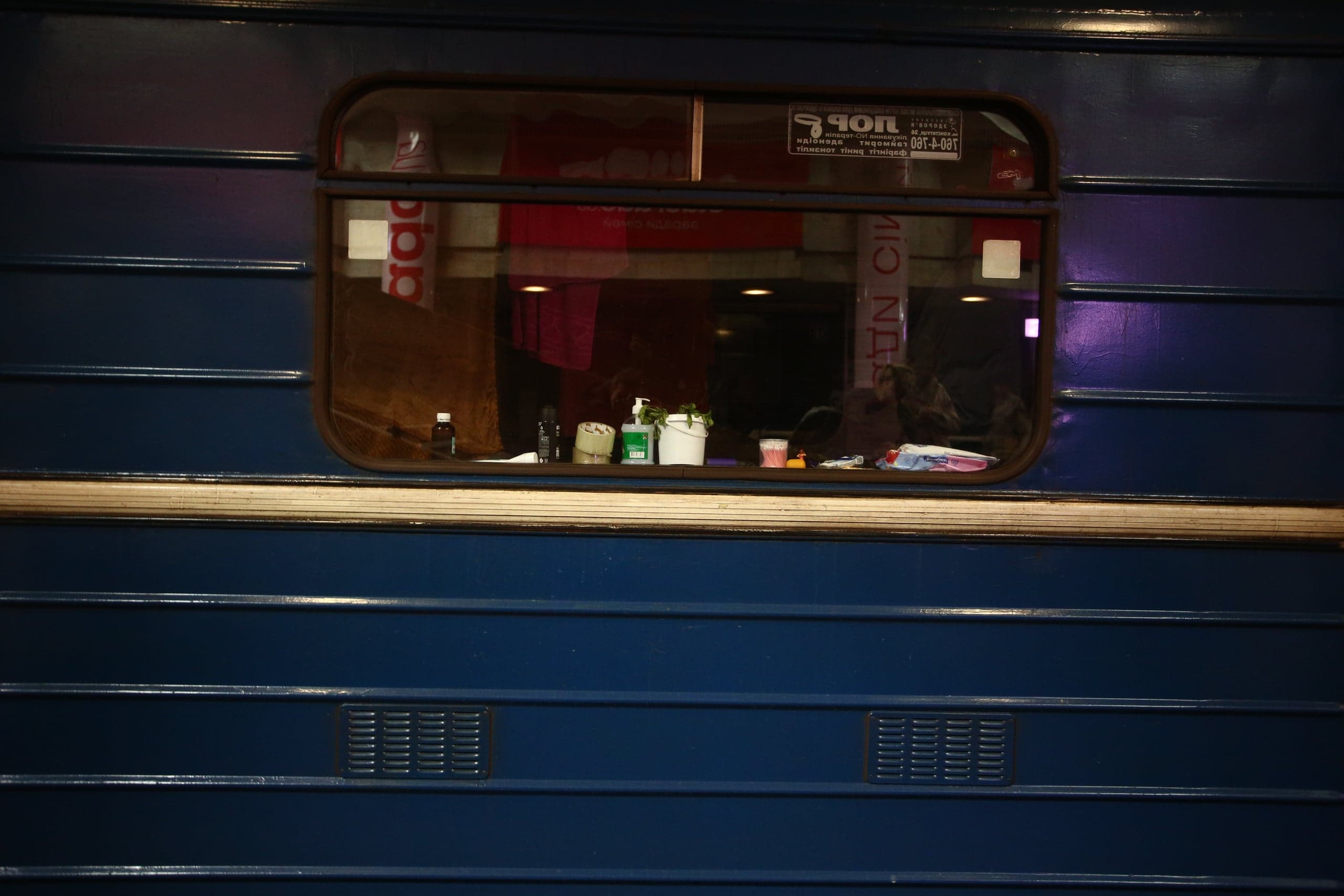 Синий ребристый вагон метро с окном, на подоконнике бытовые предметы. В отражении видны надписи "ЯОП" и "qoa".