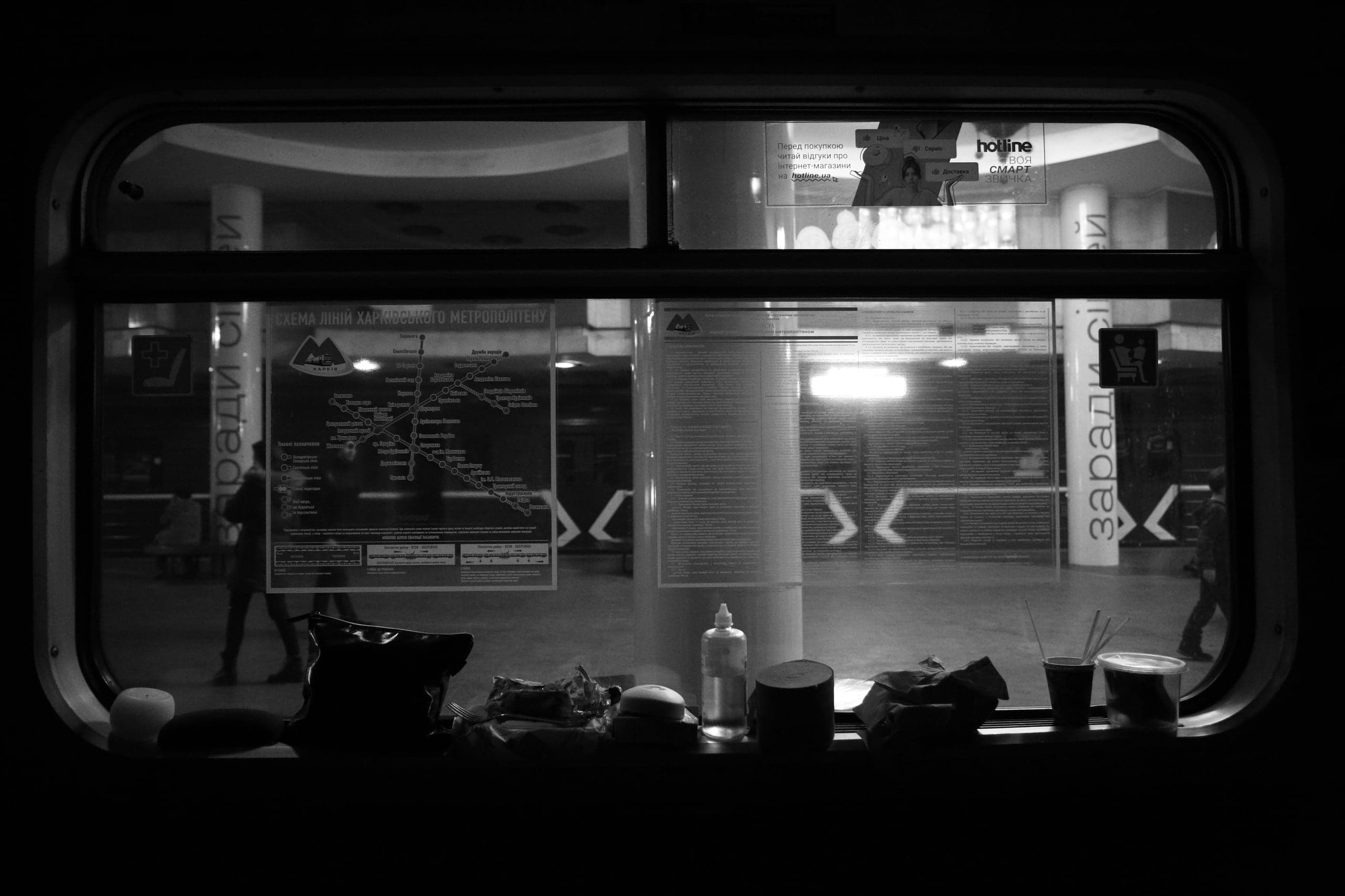 Ч/б вид из темного окна вагона метро на станцию. На подоконнике личные вещи. На платформе видна "СХЕМА ЛІНІЙ ХАРКІВСЬКОГО МЕТРОПОЛІТЕНУ" и надпись "hotline".