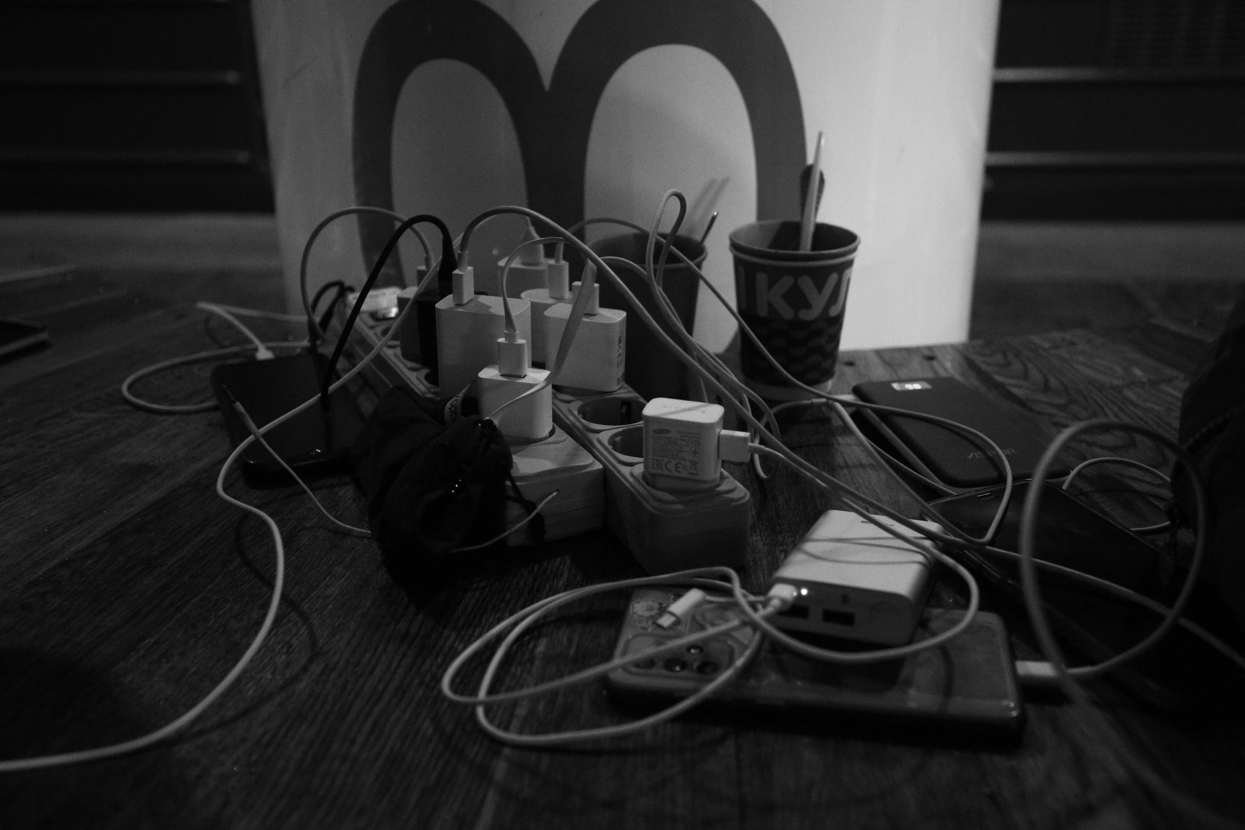 Перегруженный удлинитель, множество зарядных устройств и спутанные кабели, подключенные к телефонам и пауэрбанку на деревянном полу. На стакане виден текст "KY". Снимок монохромный.