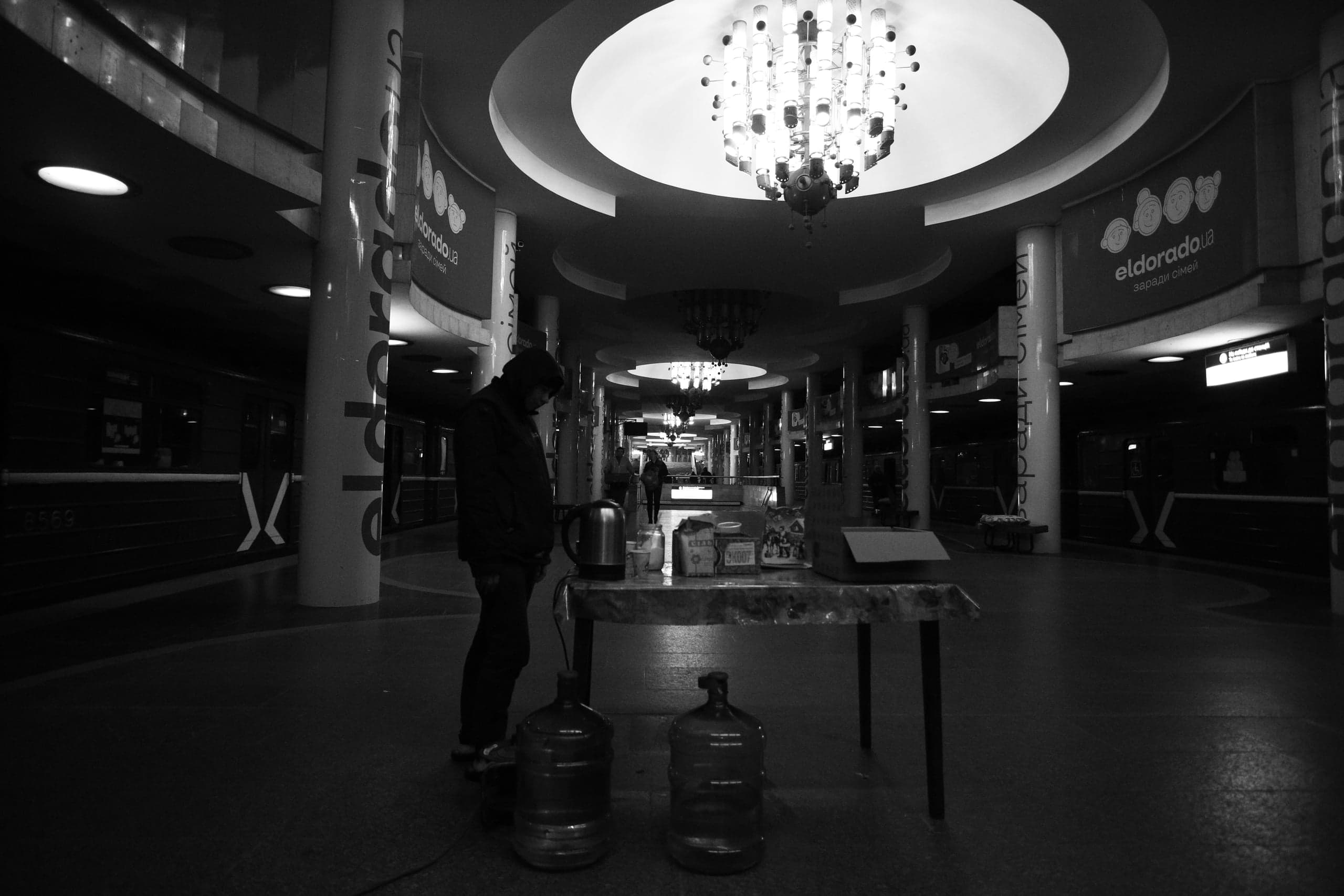 Черно-белое фото станции метро с яркой люстрой. Человек стоит у стола с чайником и бутылями воды. На фоне реклама: "eldorado.ua", "заради сімей".