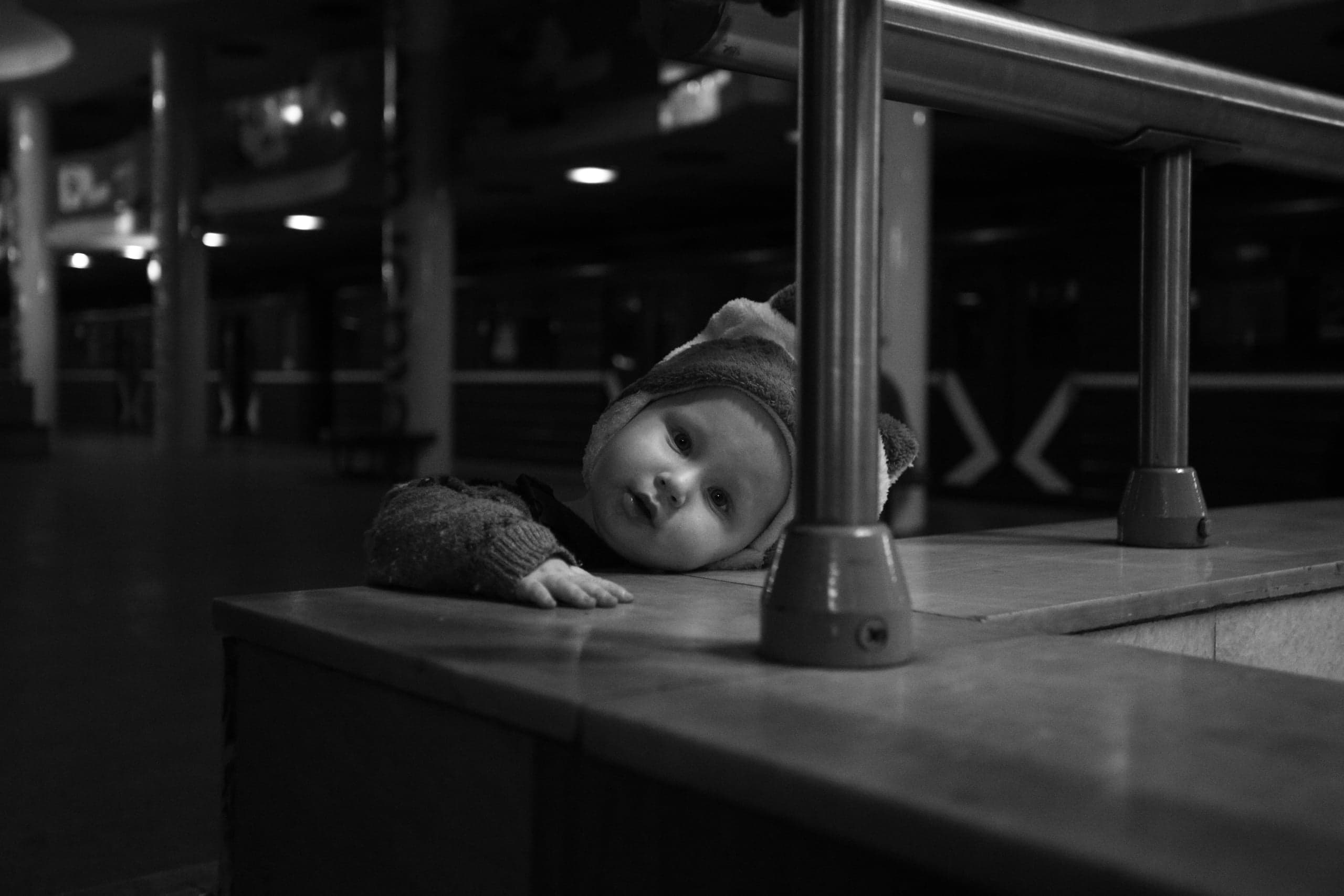 Монохромный снимок: младенец в шапке, лежащий на низком парапете, смотрит в кадр, обрамленный металлической стойкой на темном фоне.
