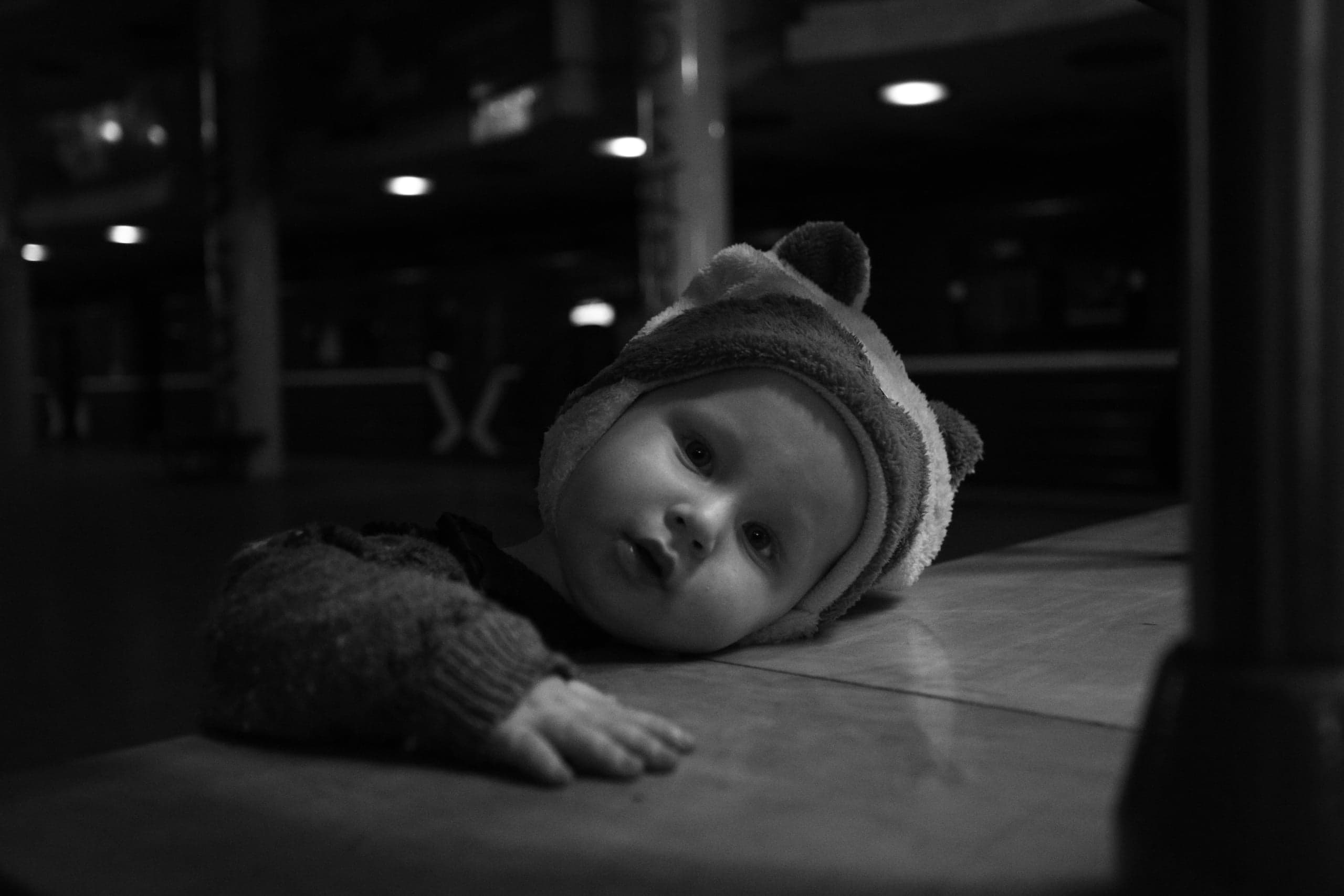Черно-белое фото: младенец в шапке лежит на столе, вглядываясь в камеру на фоне темного интерьера.