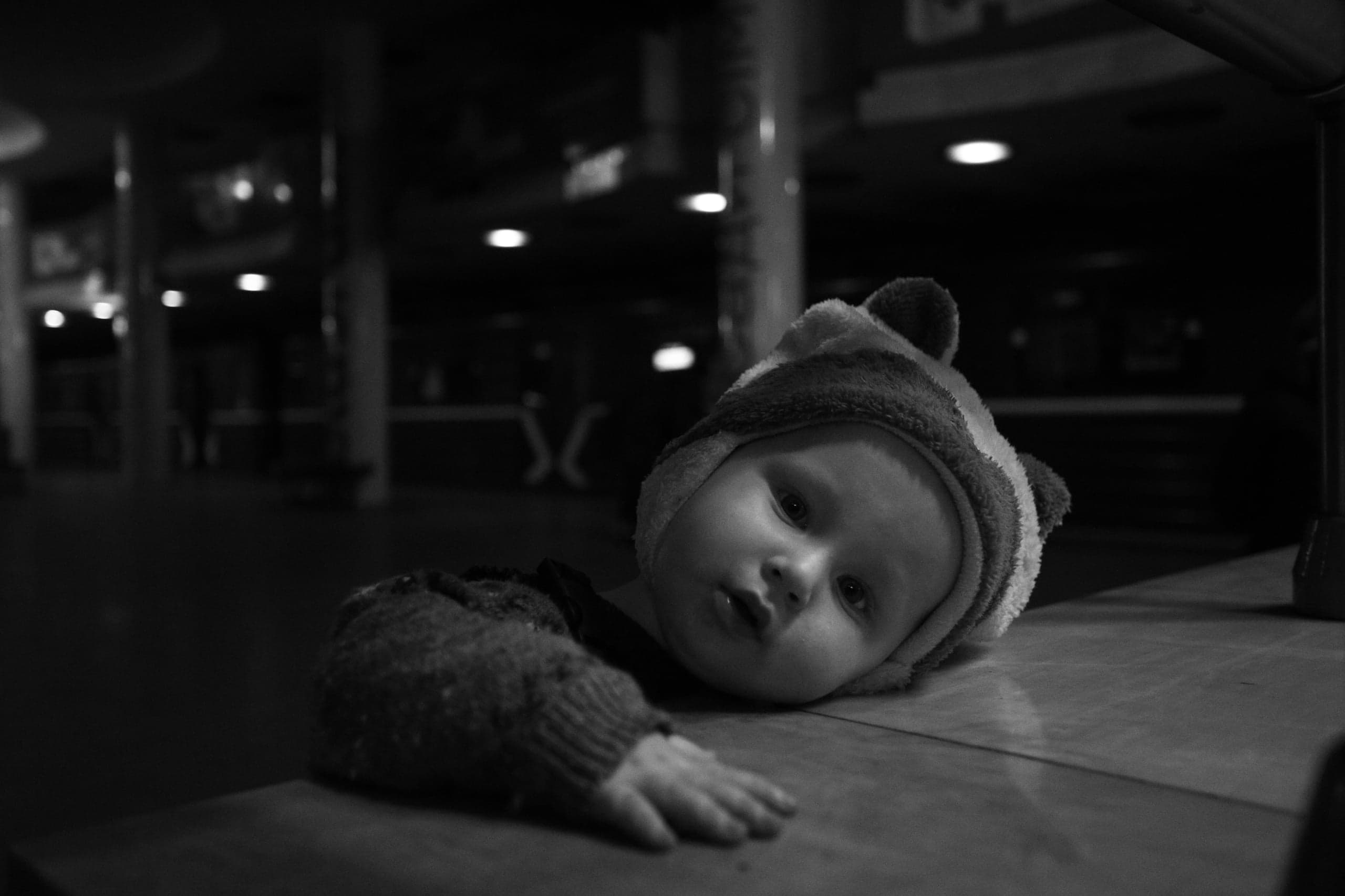 Монохромный снимок: ребенок в шапке, лежащий на столешнице на фоне темного, плохо освещенного зала.
