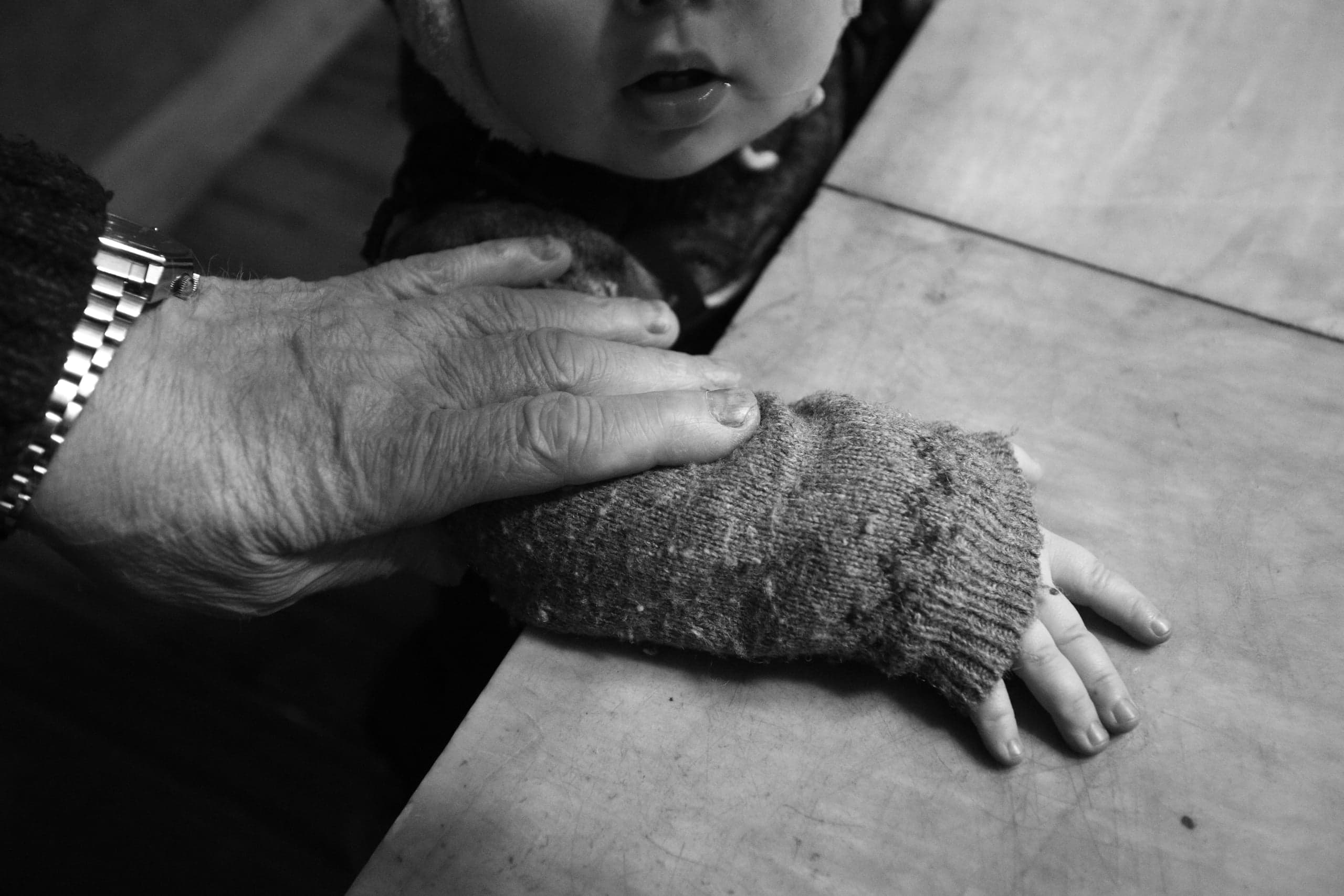 Чёрно-белое фото: рука пожилого человека с часами на руке ребенка в вязаном рукаве. Видна нижняя часть лица ребенка.