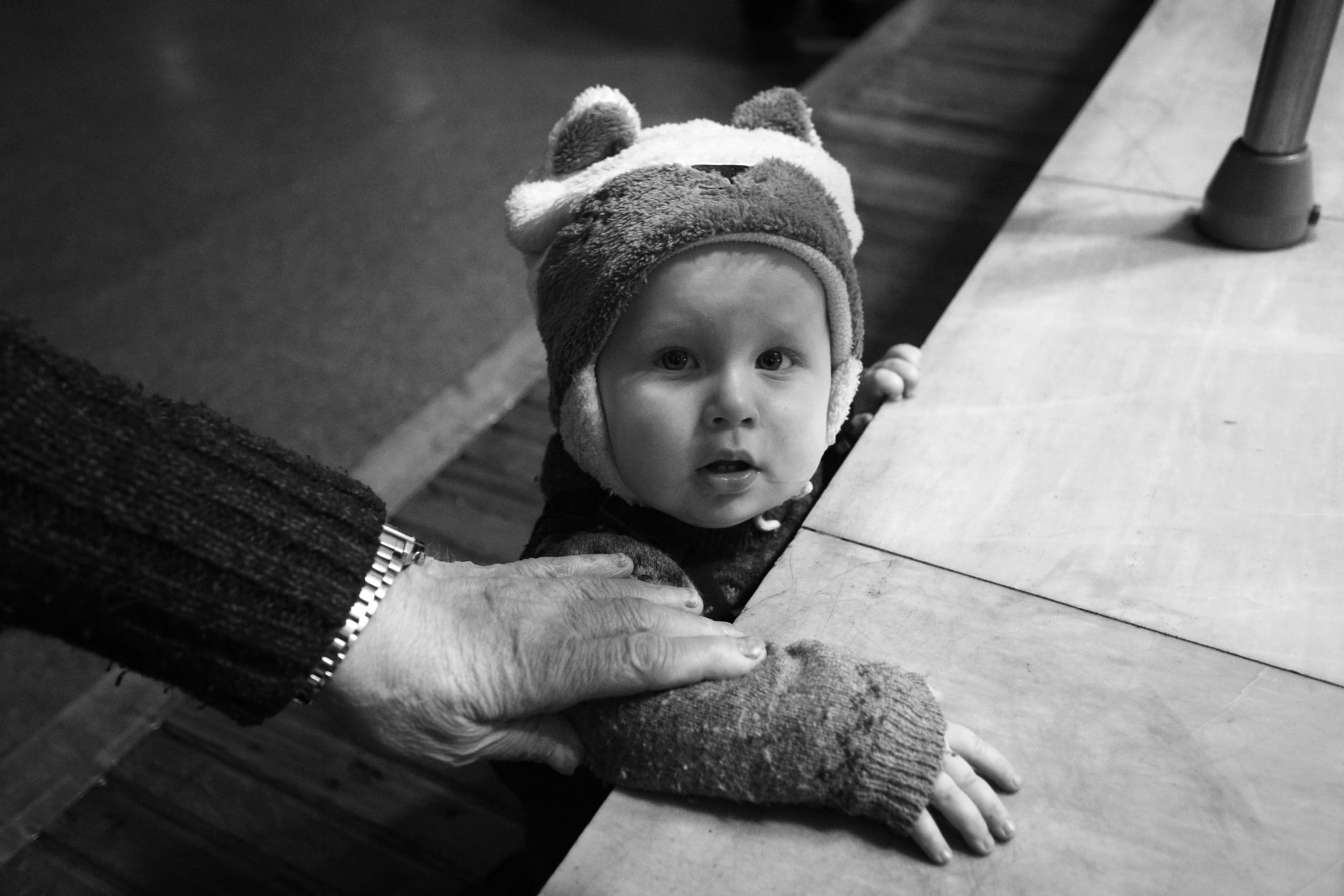 Черно-белый снимок: ребенок в шапке с ушками выглядывает из-за стола, пожилая рука с часами лежит на его предплечье.