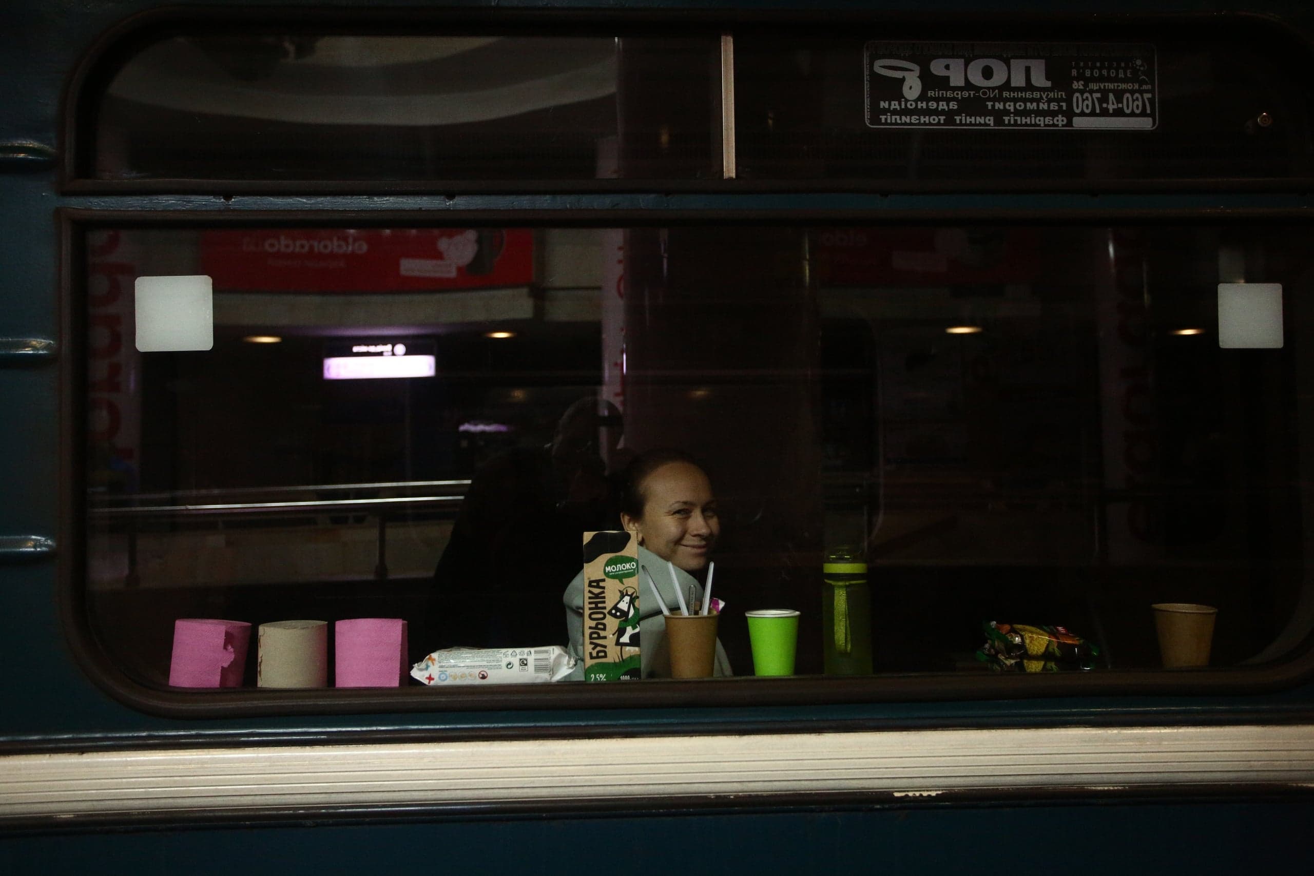 Улыбающаяся женщина за окном вагона метро. На подоконнике разложены: пакет молока "БУРЬОНКА", стаканы и рулоны бумаги.