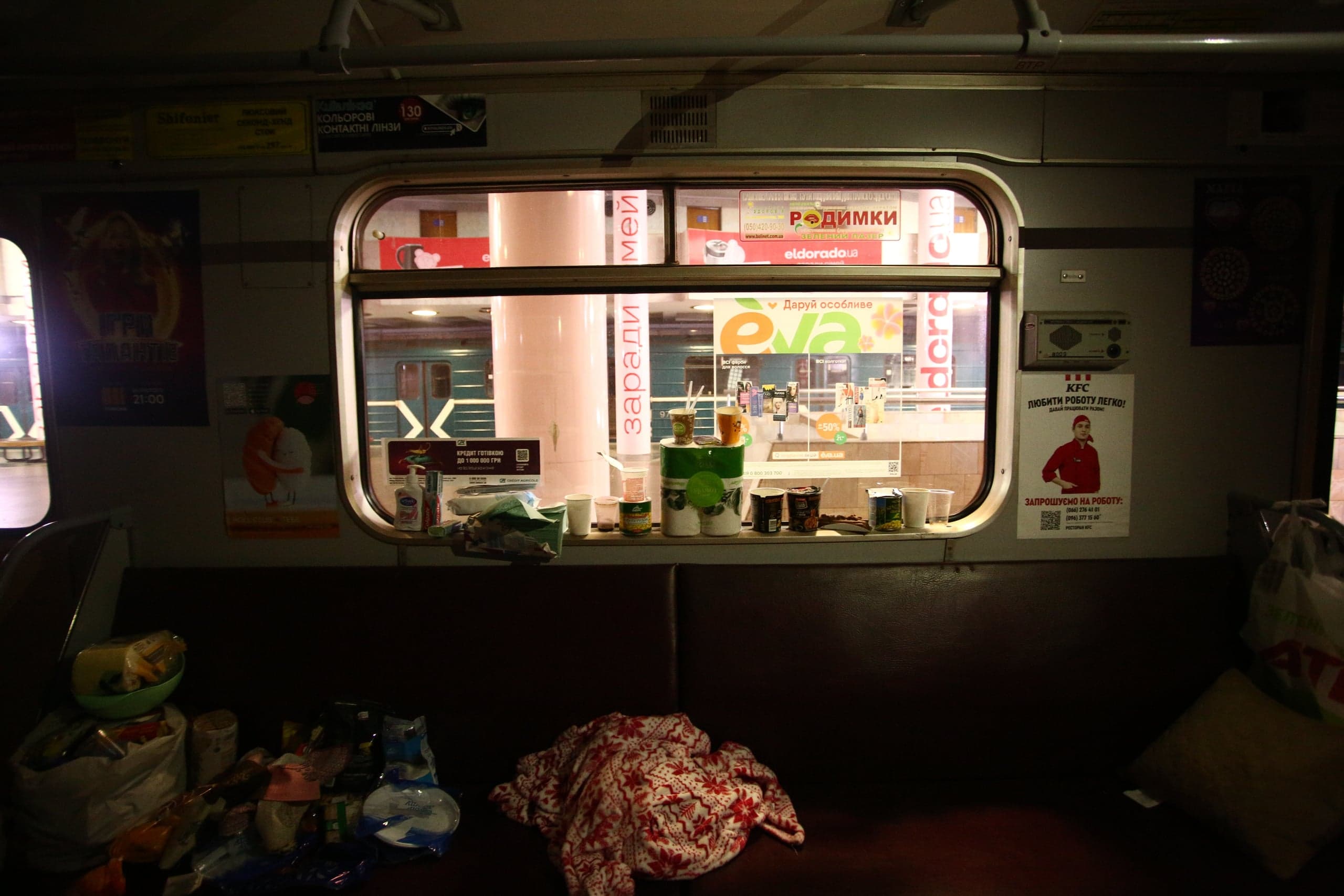 Темный интерьер вагона метро, сиденья завалены личными вещами, сумками и одеялом. На подоконнике стоят чашки и контейнеры. В окне видна ярко освещенная станция с рекламой "eva" и "eldorado.ua". На правой стене плакат "KFC: ЛЮБИТИ РОБОТУ ЛЕГКО! ЗАПРОШУЮ НА РОБОТУ:".