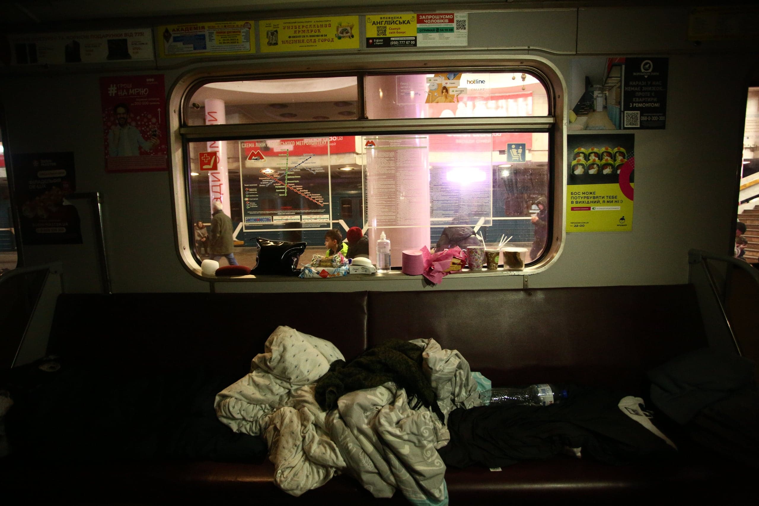 Интерьер темного вагона метро, припаркованного на ярко освещенной станции. На сиденье свалены одеяла и личные вещи. В окне видна карта "СХЕМА ЛІНІЙ".