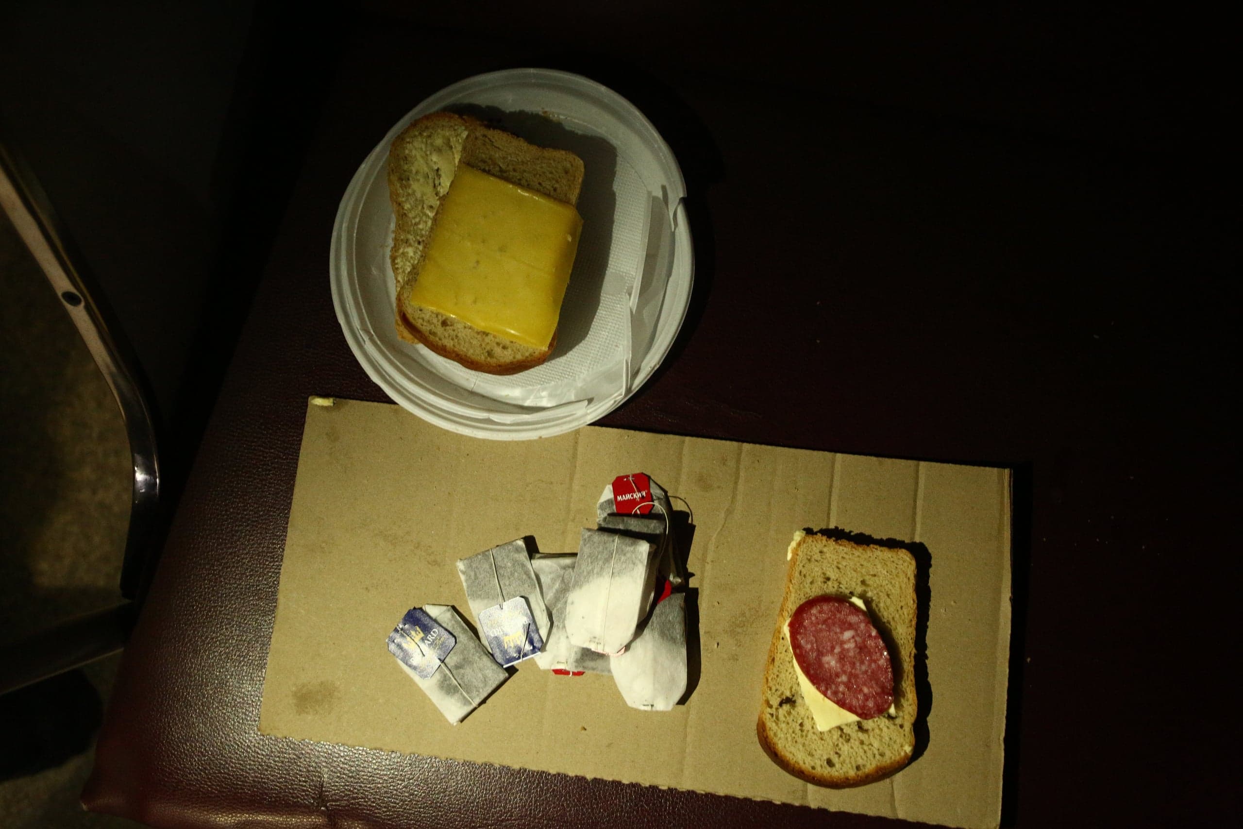 Сэндвич с сыром на пластиковой тарелке, сэндвич с колбасой и несколько чайных пакетиков на куске картона. Освещение приглушенное.