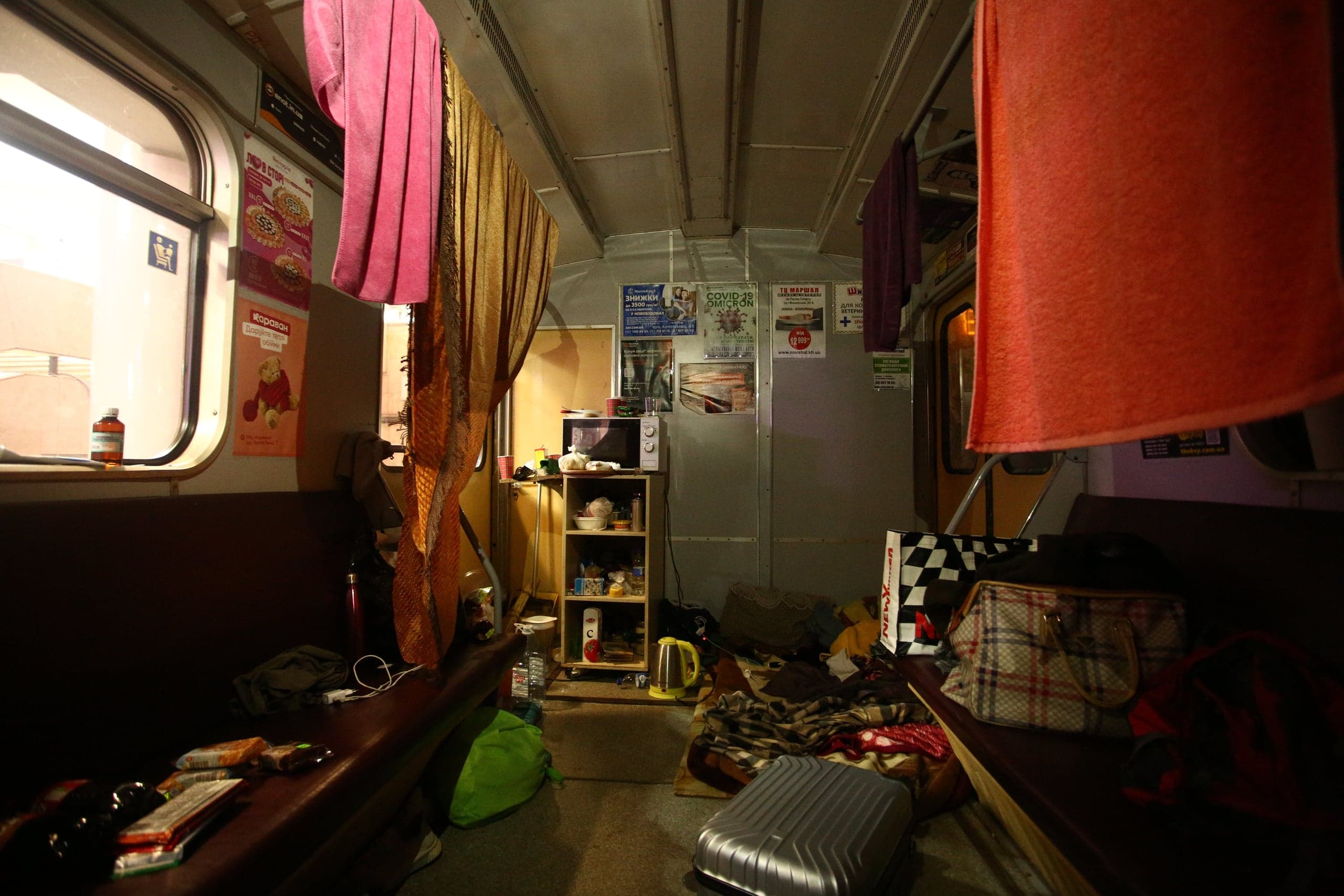 Переполненный вагон поезда, переоборудованный в жилое помещение, с полками, микроволновой печью и висящими полотенцами. На стенах плакаты: "COVID-19 Omicron".