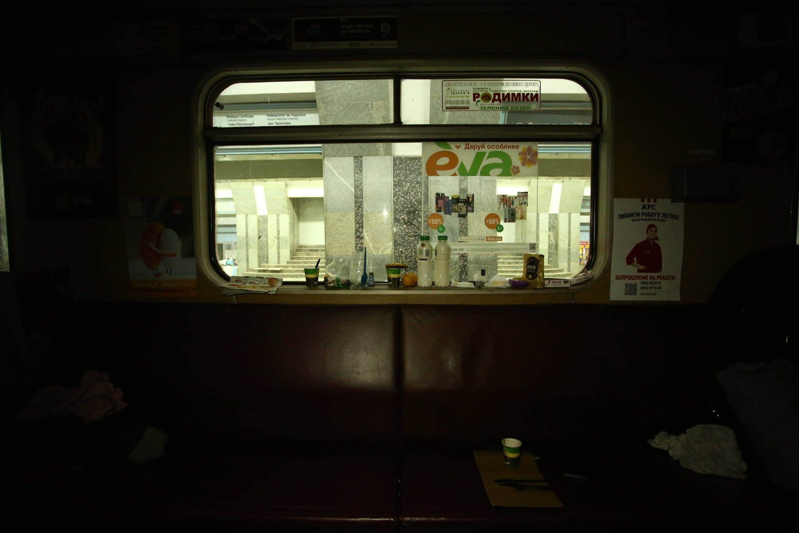 Темный интерьер (возможно, киоск) с красными сиденьями. Через окно виден ярко освещенный вход в метро с мраморными лестницами. Подоконник заставлен бутылками и мусором. На стенах видны постеры "EVA" и "KFC ЛЮБИТИ РОБОТУ ЛЕГКО!".