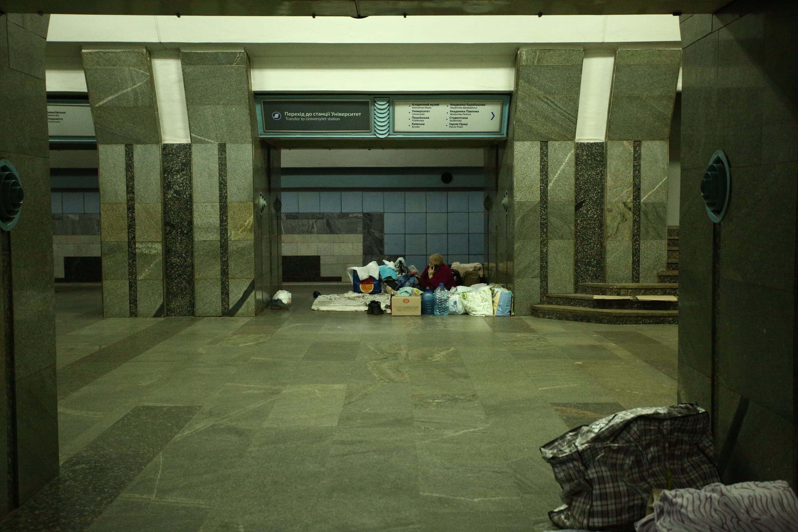 Вестибюль метро с гранитными колоннами. Человек сидит среди личных вещей и запасов воды, используя станцию как убежище. На знаке: "Перехід до станції Університет".