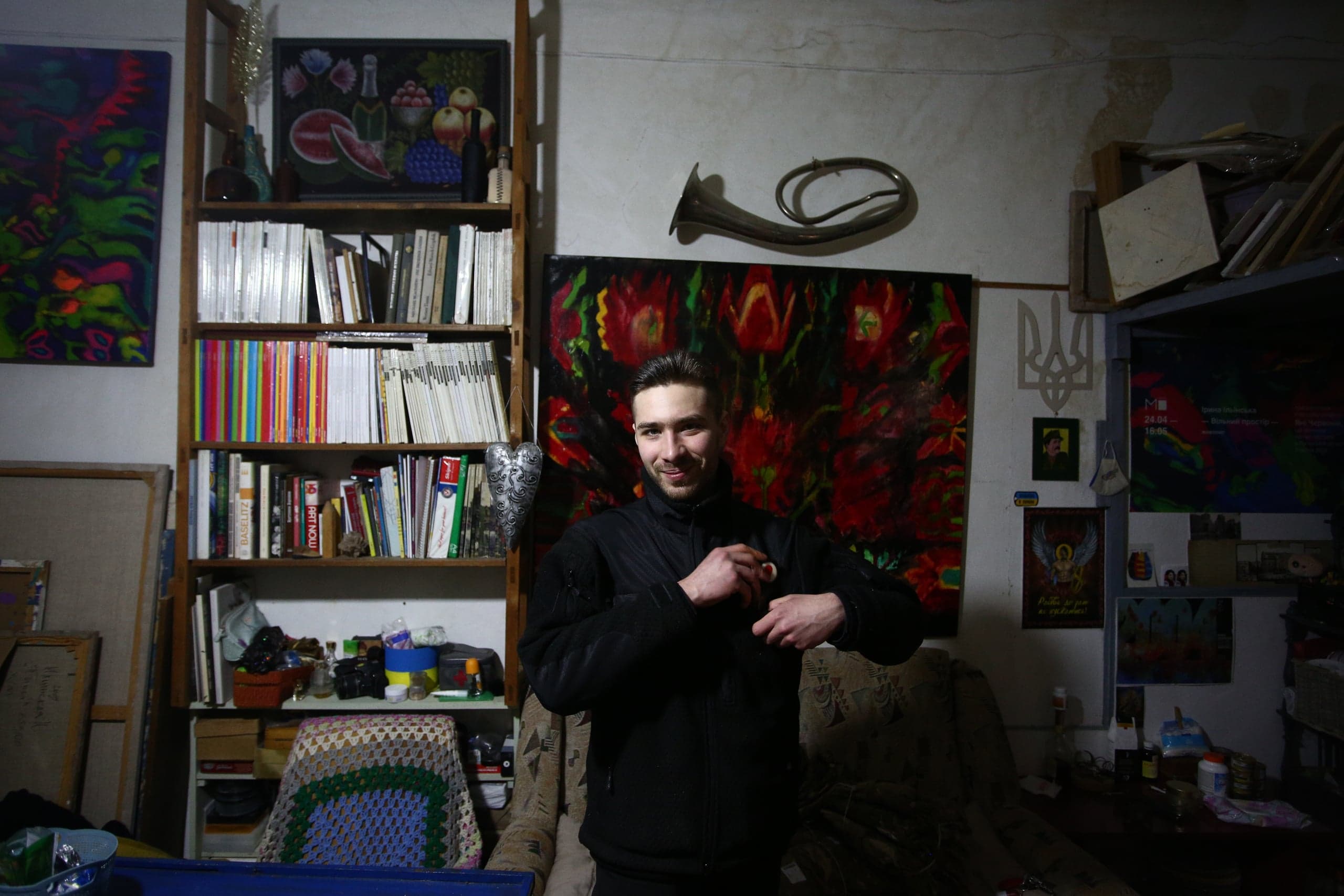 Улыбающийся мужчина в студии. На фоне: книжный шкаф, большая картина с красными цветами, горн и украинский Тризуб.