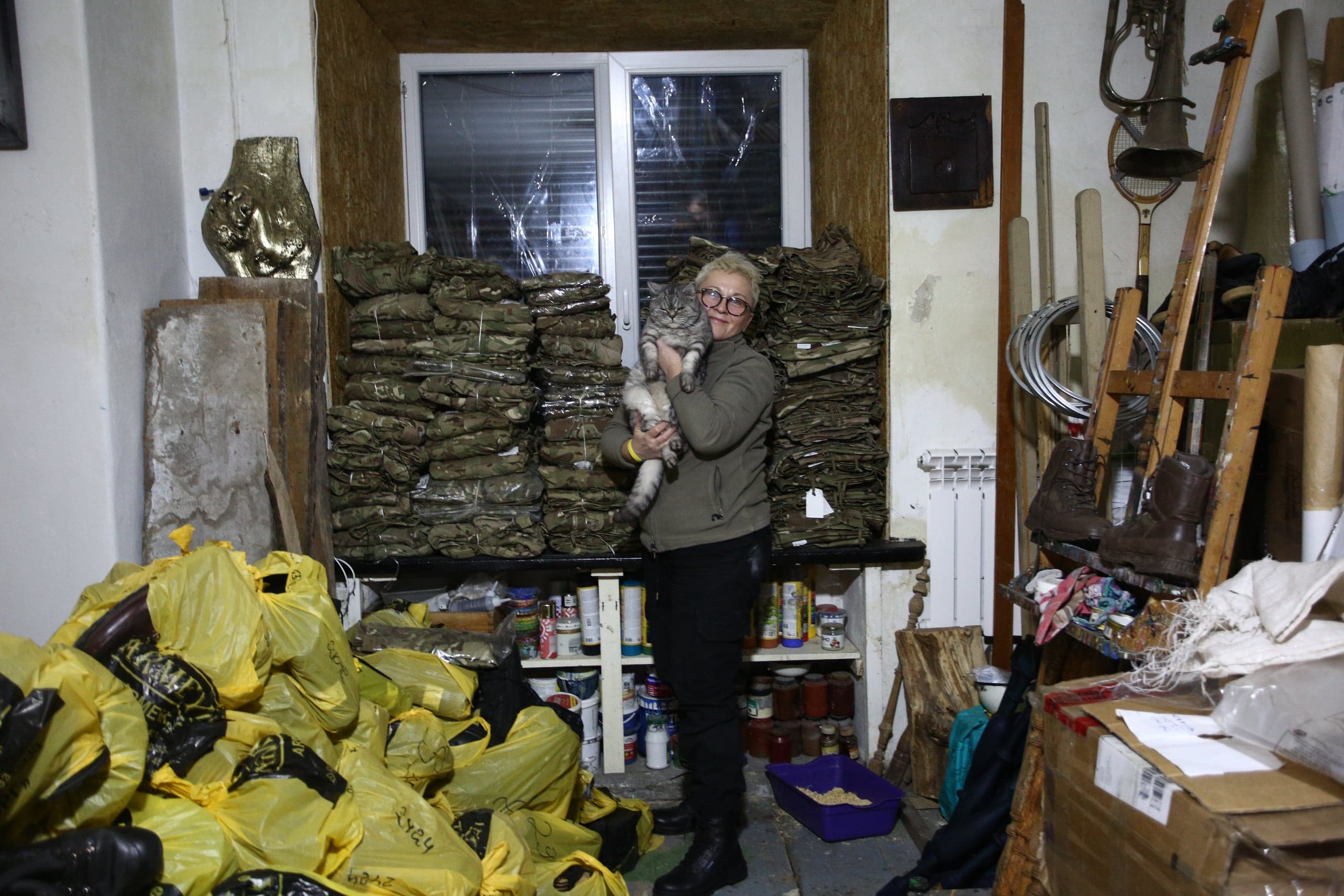 Женщина в очках держит кота, стоя в комнате, заваленной желтыми мешками с гуманитарной помощью и стопками камуфляжной ткани. Стены обставлены разнородными предметами и инвентарем.