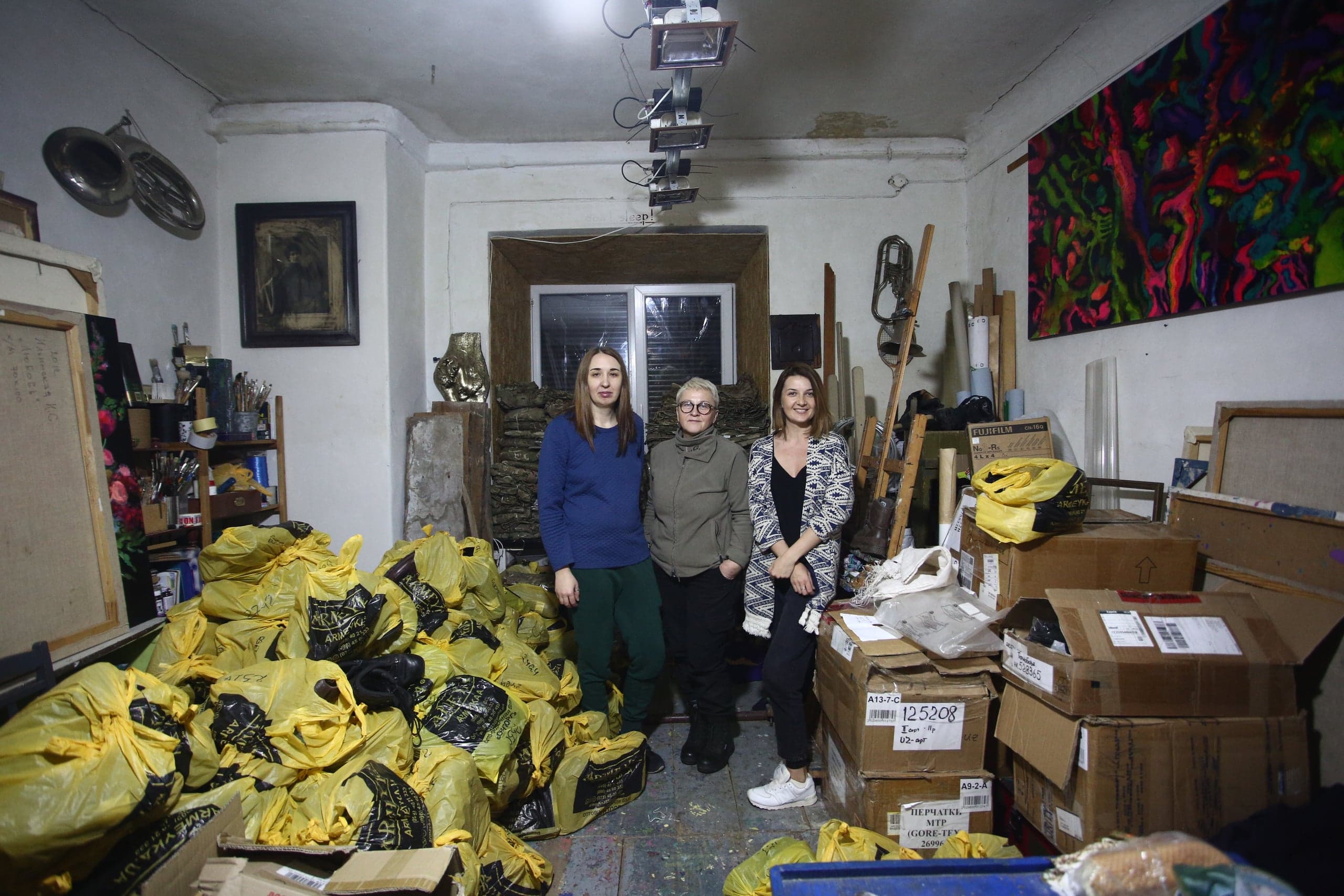 Три женщины стоят в студии среди горы желтых мешков и стопок картонных коробок.