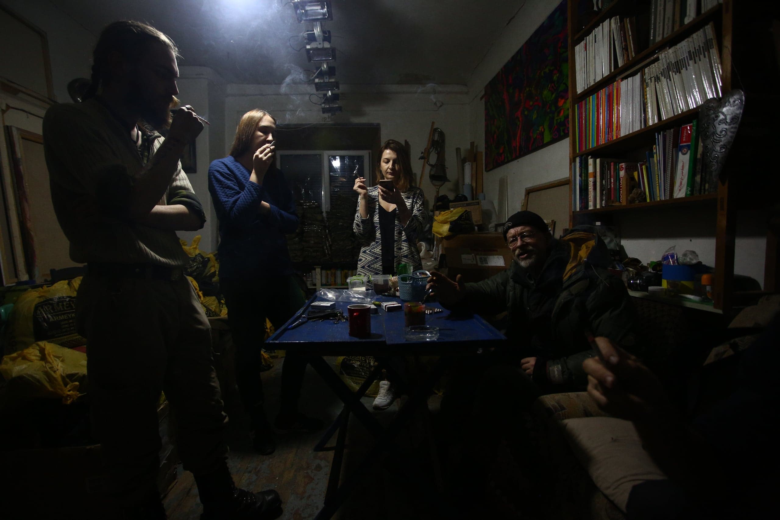Пятеро людей отдыхают и курят в тускло освещенной комнате вокруг стола. Справа — книжный шкаф, слева сложены желтые мешки.