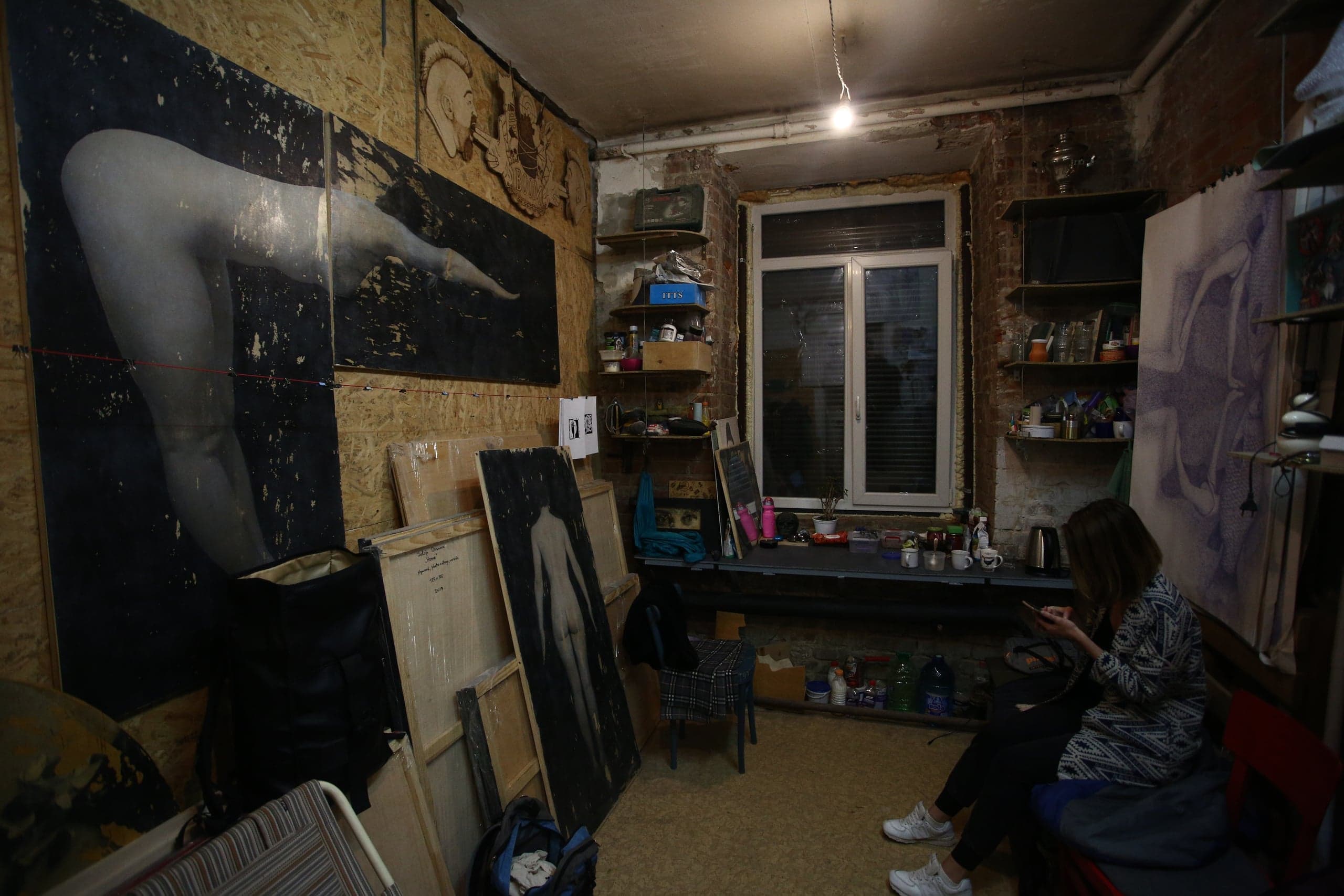 Художественная студия. Стены обшиты фанерой, на них висят крупные черно-белые картины обнаженной натуры. Женщина сидит справа, глядя в телефон.