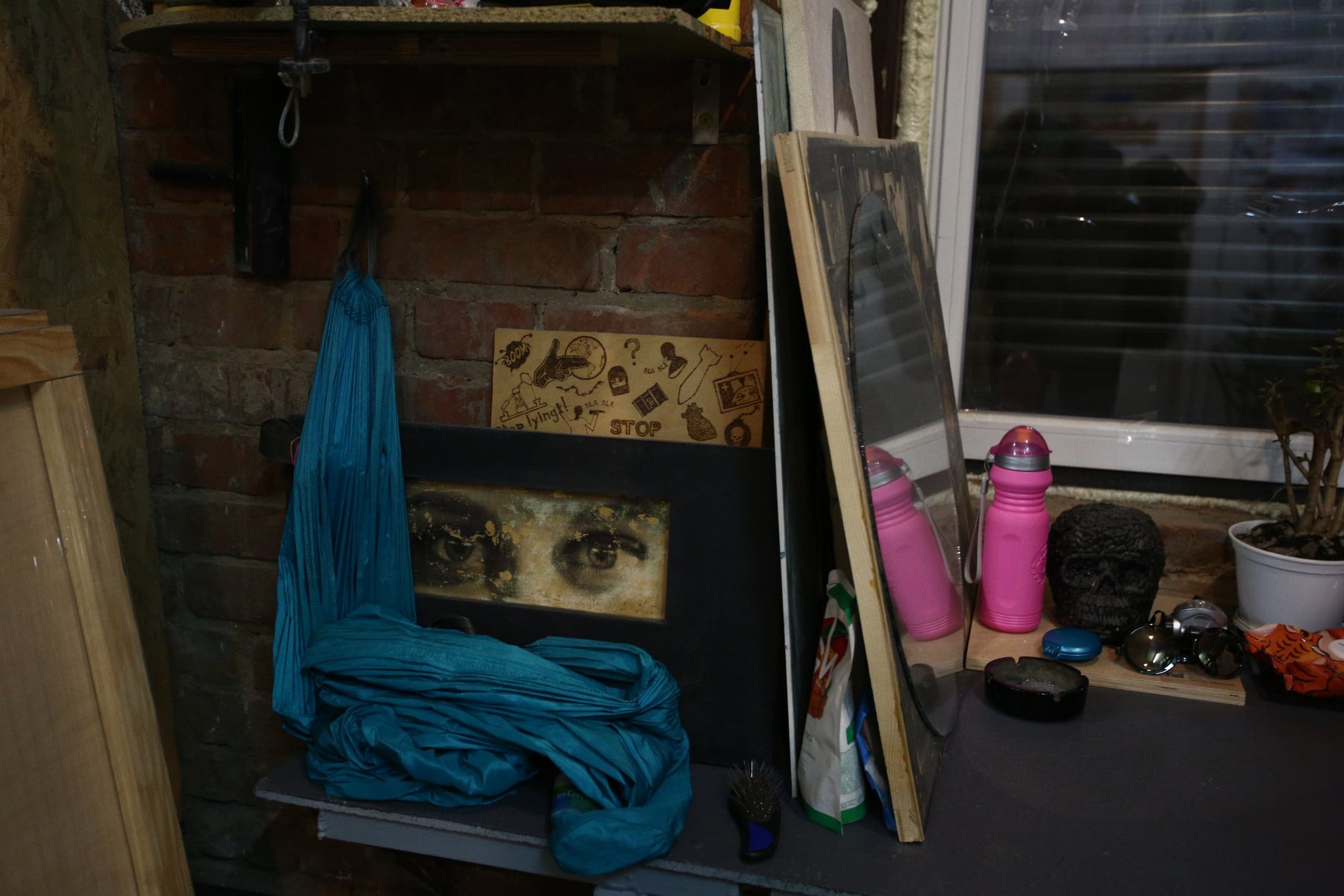 Уголок у кирпичной стены, освещенный окном. На полке лежат синяя ткань и рамка с изображением глаз. На дощечке текст: «lying!» и «STOP». На столе стоят две розовые бутылки, черный декоративный череп и зеркало.