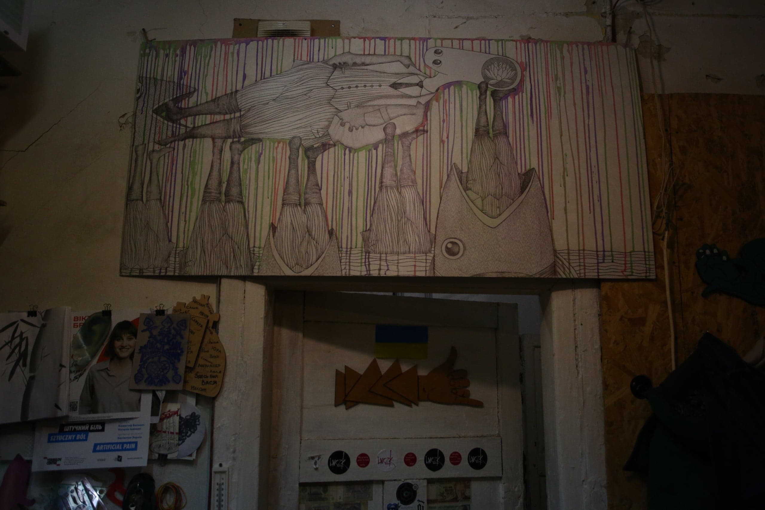 Сюрреалистический графический рисунок над дверным проемом в темном помещении: перевернутая фигура на конусообразных столбах на фоне цветных потеков. Под дверью: украинский флаг и постеры, включая "ШТУЧНИЙ БІЛЬ" и "ARTIFICIAL PAIN".