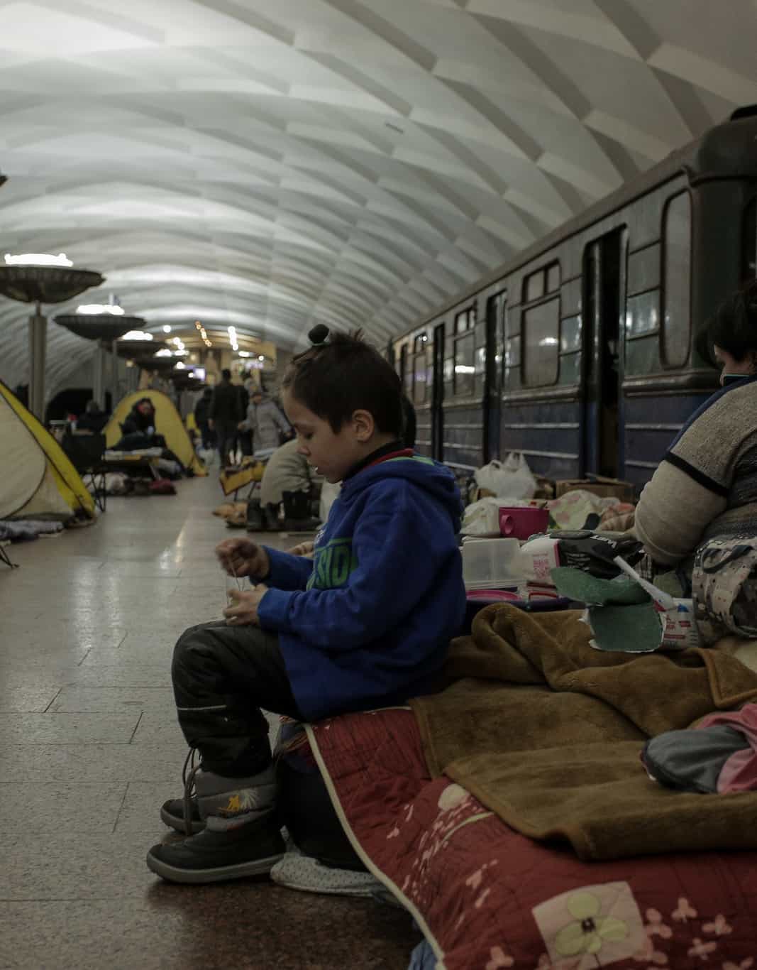 A child sitting on a mattress in a subway station that is being used as a bomb shelter.